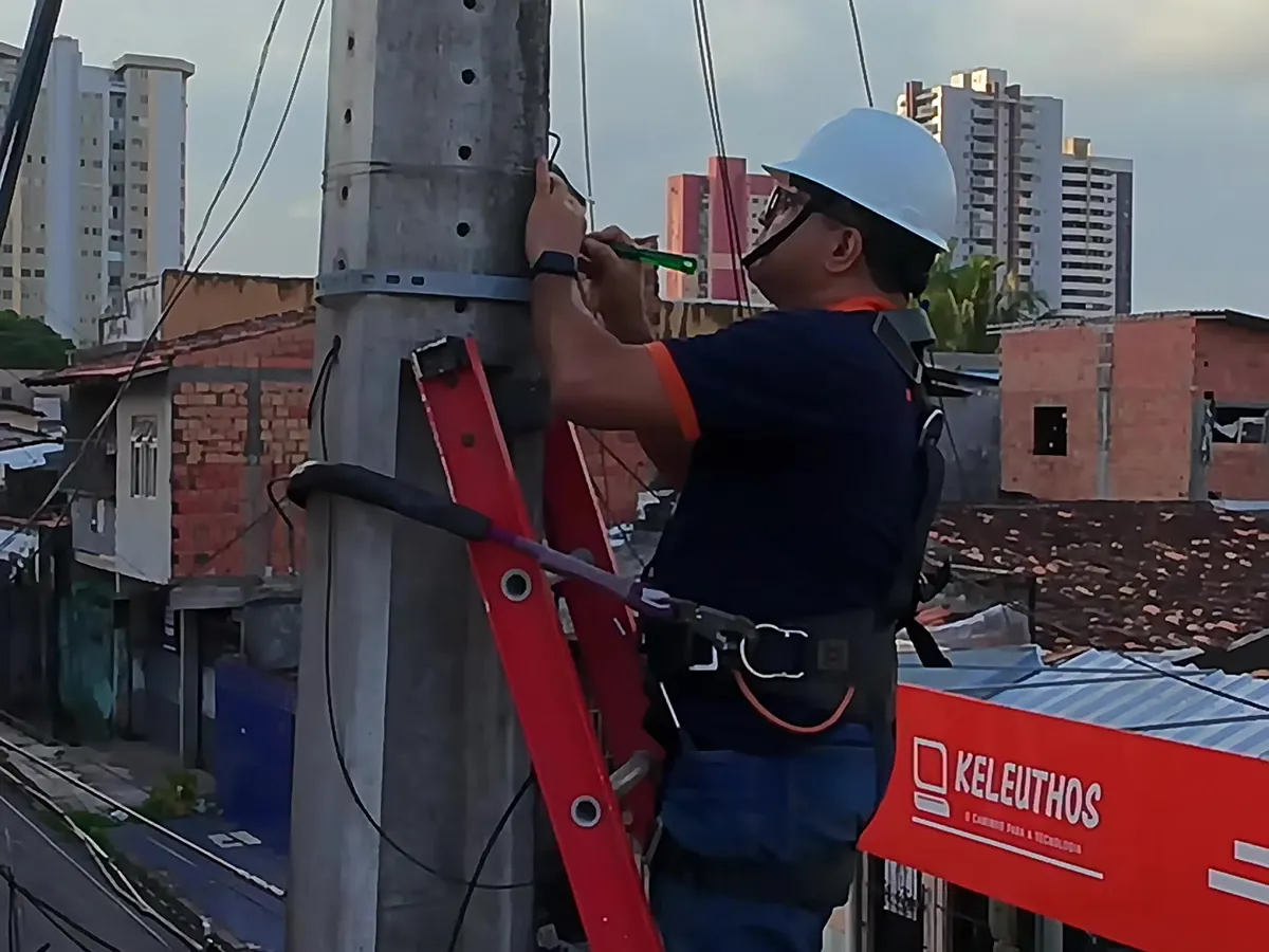 Técnico com capacete e cinto de segurança no alto da escada, instalando a câmera nova em frente à fachada da Keleuthos